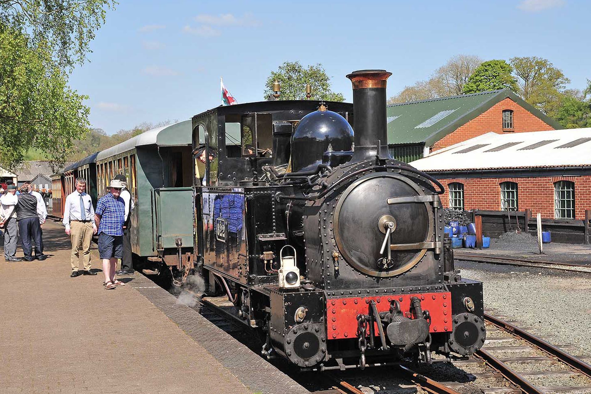 Black steam locomotive at a station platform with a few people nearby and a red-brick building in the background