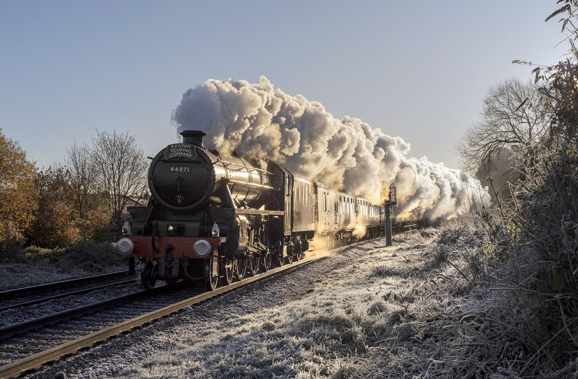 Steam locomotive pulling cars through snowy countryside at dusk, with thick white steam billowing behind.