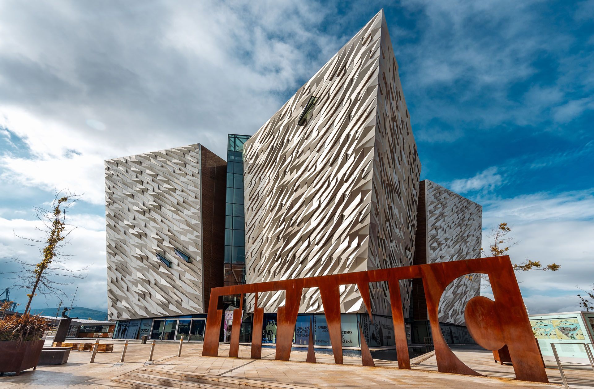 Modern angular buildings beside a rust-red sculpture under a cloudy blue sky