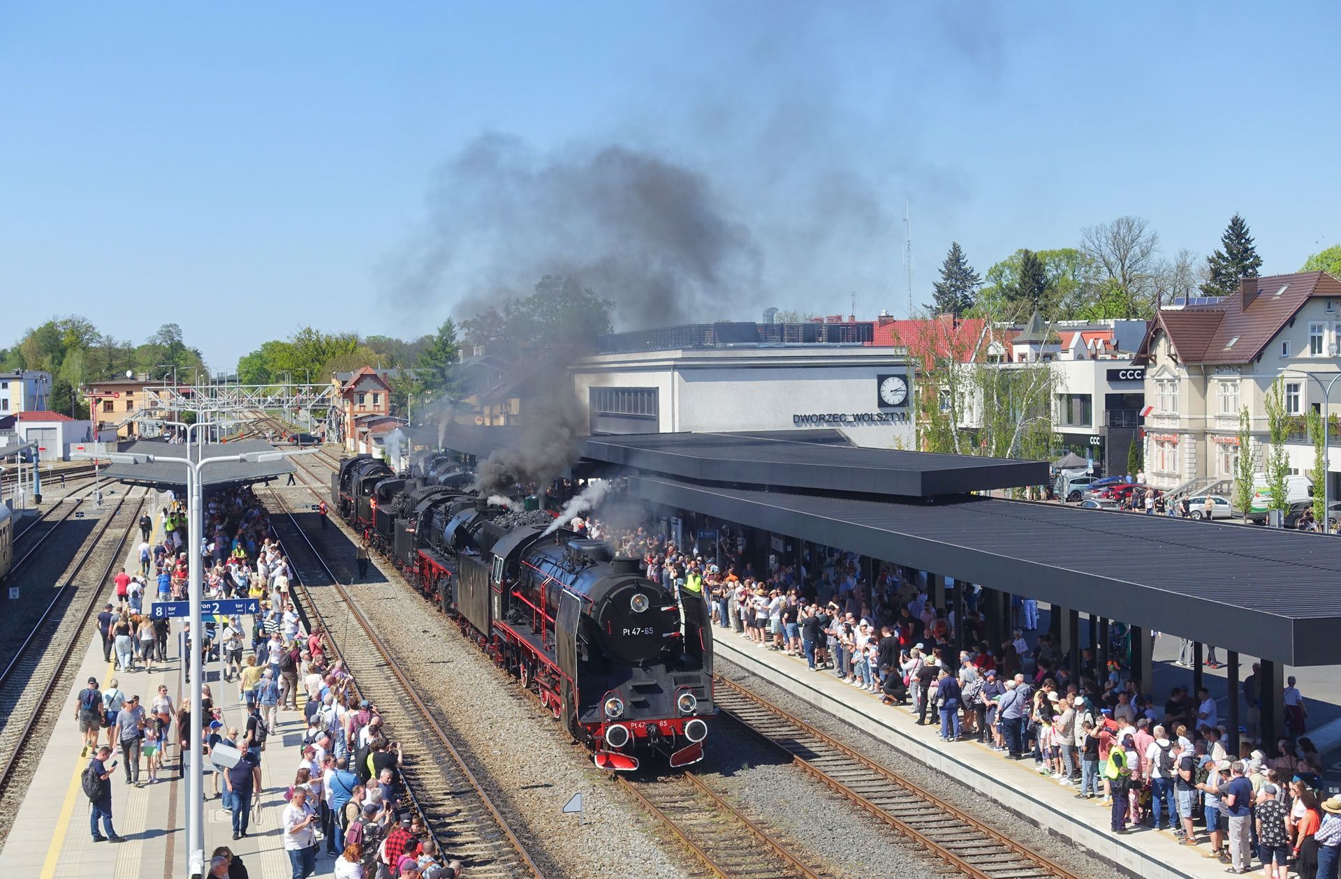 Steam locomotive at a crowded station platform, with smoke rising as passengers watch nearby.