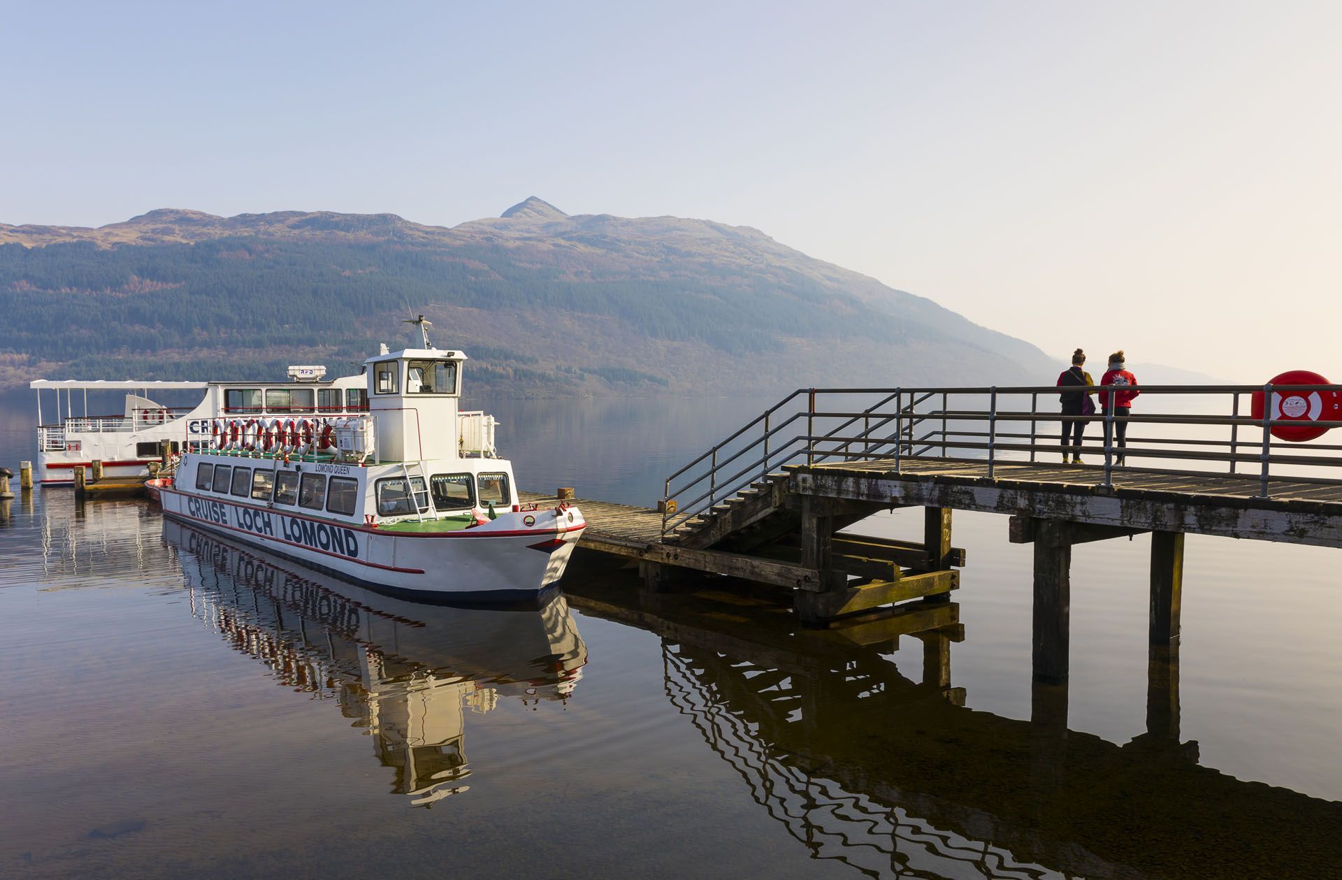 Passenger boat docked at a lake pier with two people standing on the ramp, mountains in the background