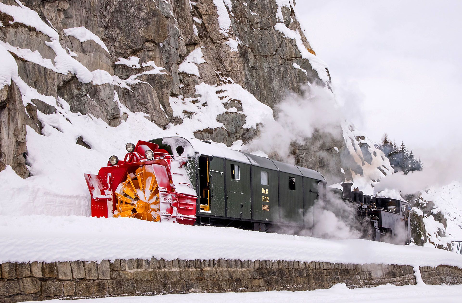Steam locomotive with red snowplow pushing through deep snow beside a rocky mountain track
