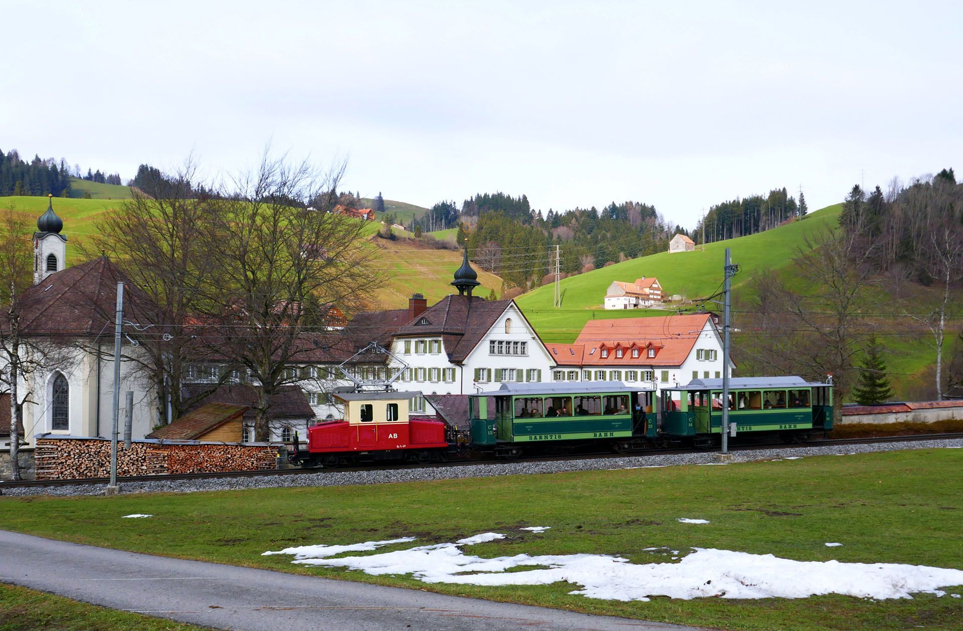 Small red and green train by white houses on a grassy hillside with patches of snow