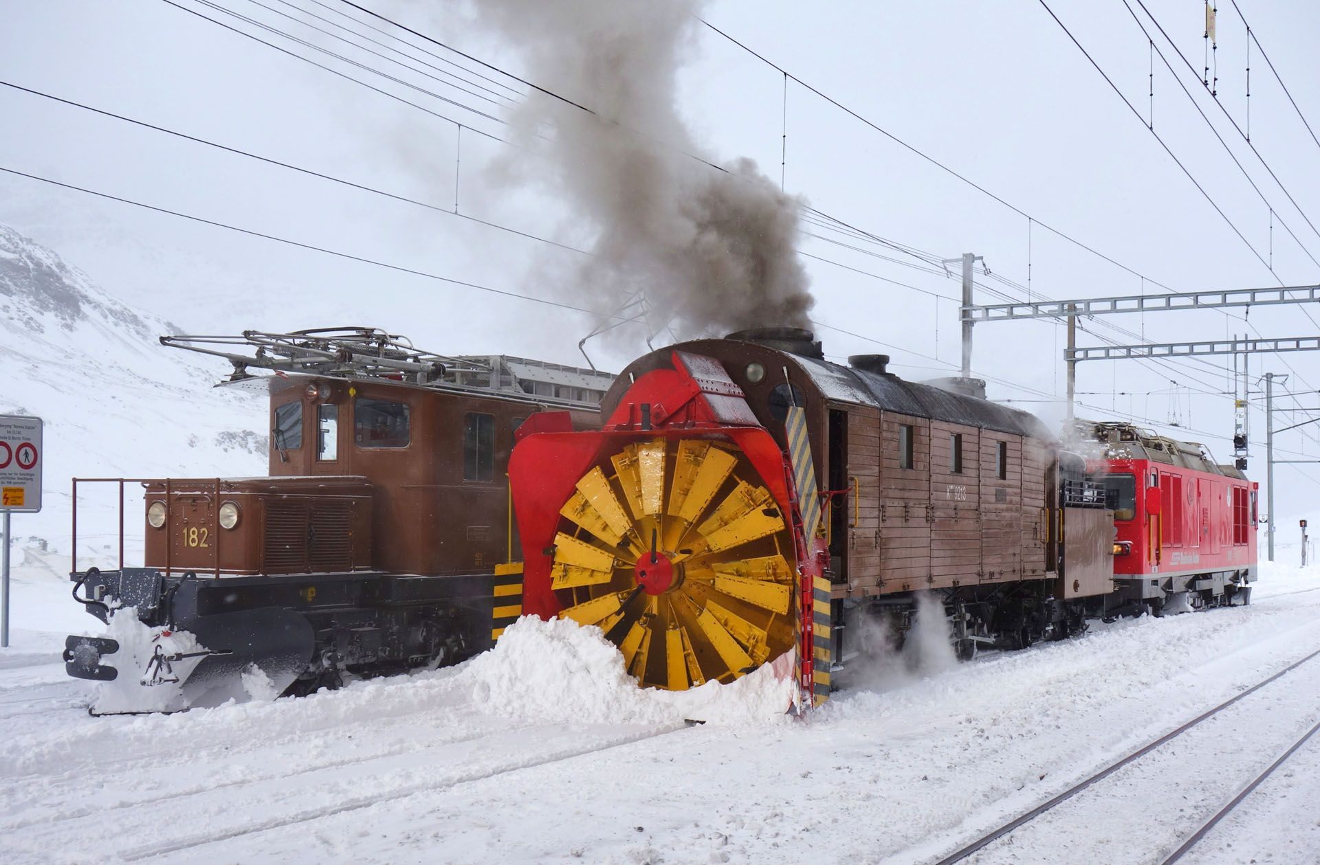 Snowplow train clearing snowy tracks, with a large yellow plow and smoke rising from the engine.