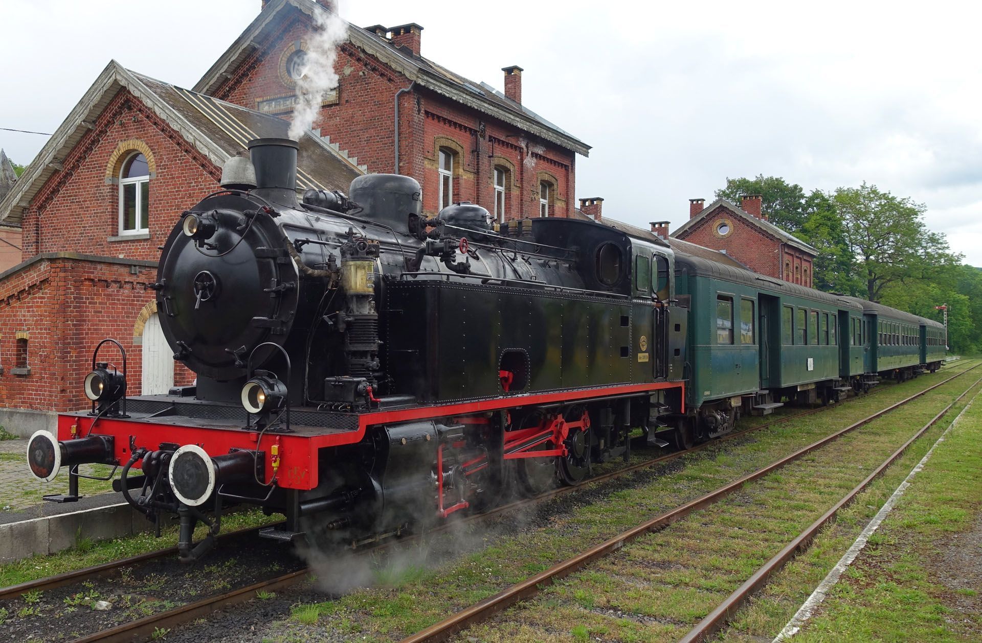Black steam locomotive pulling green passenger cars beside a brick station on rainy tracks