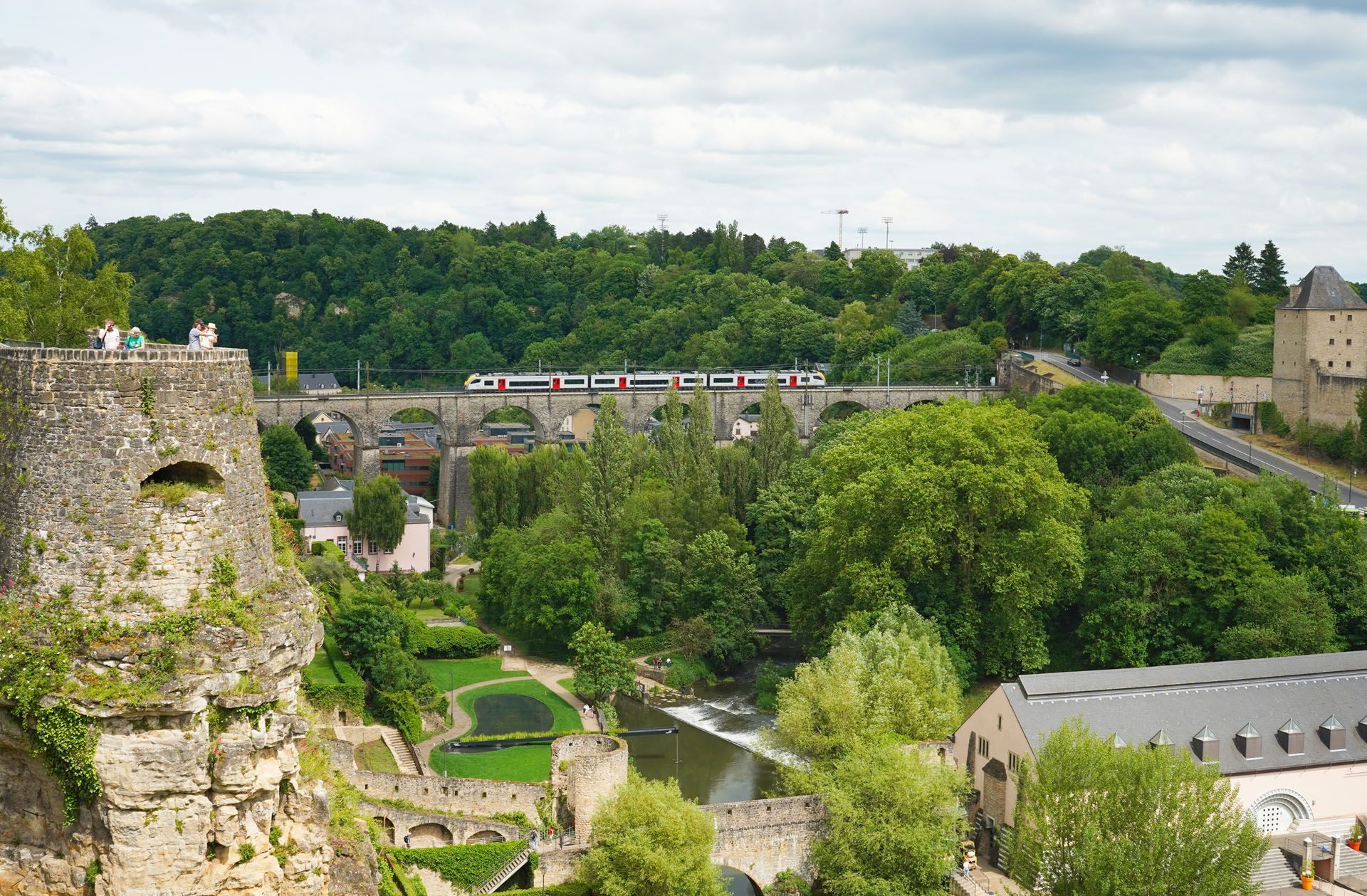 Stone ruins overlooking a lush green valley, a river, and a stone bridge under a cloudy sky.
