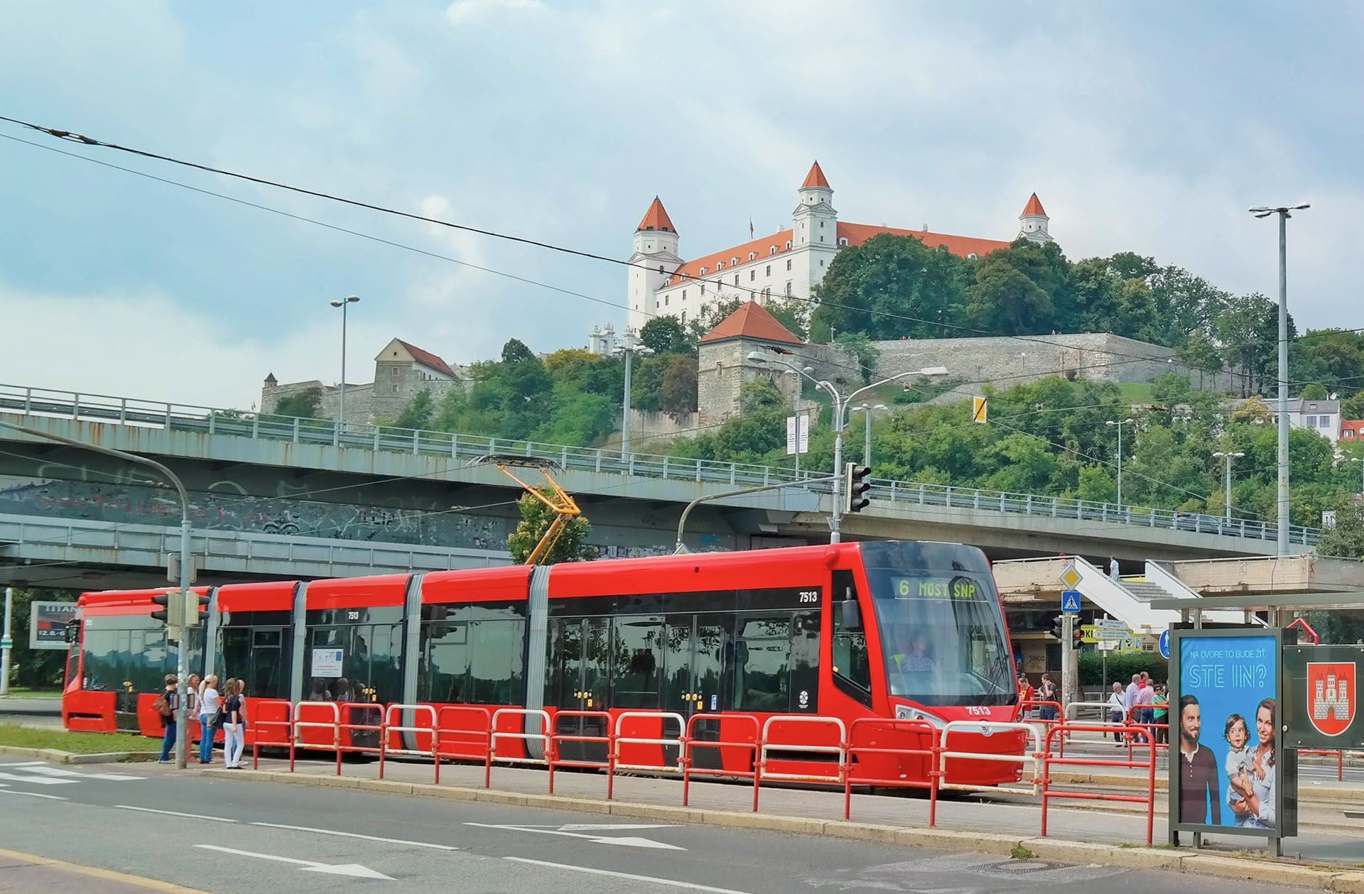Red tram at a stop in front of a hilltop castle with red roofs and a cloudy sky