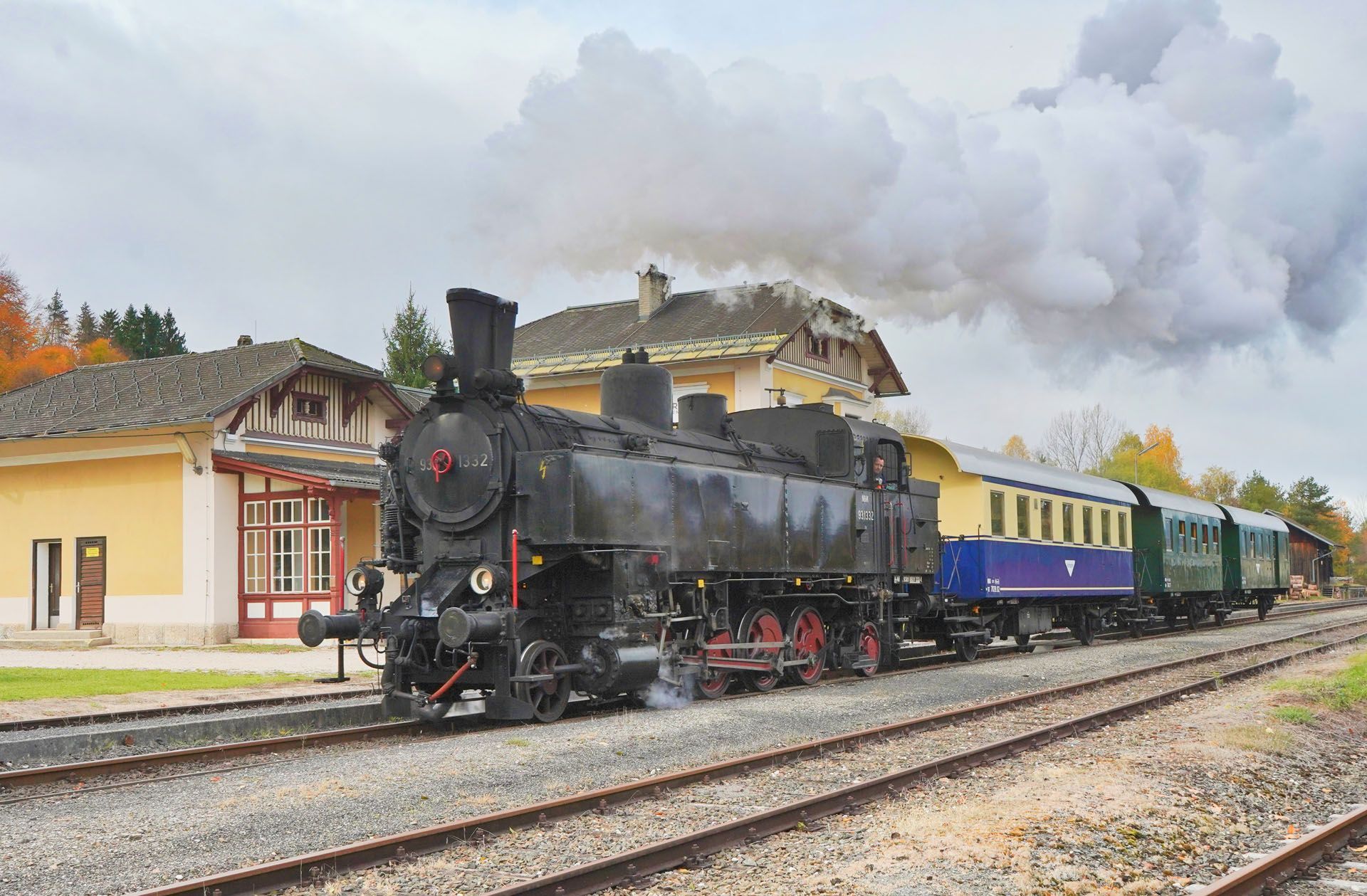 Black steam locomotive pulling colorful passenger cars past a station under billowing smoke.