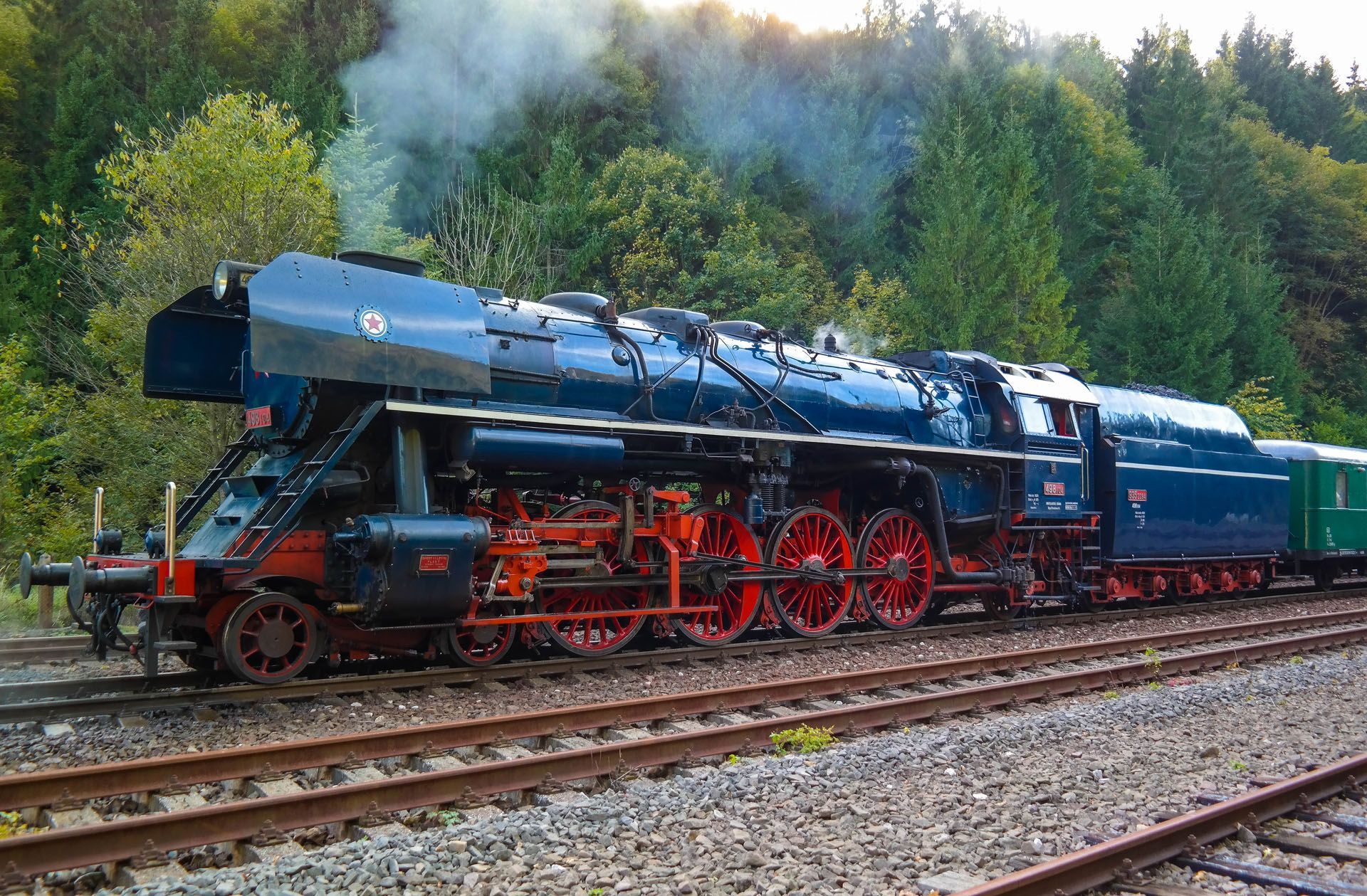Black steam locomotive with red wheels on tracks, billowing smoke in a wooded setting
