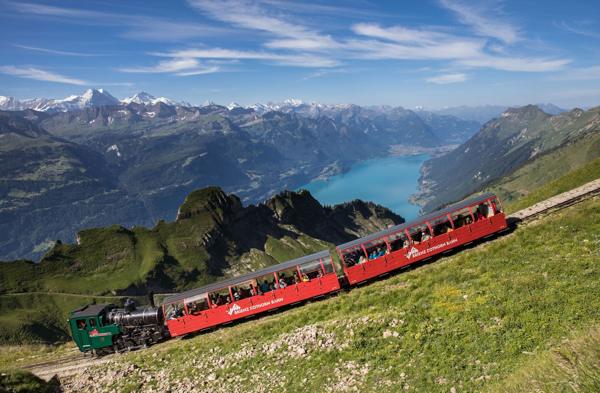 Red mountain train climbing a grassy slope above a blue lake and snowy alpine peaks