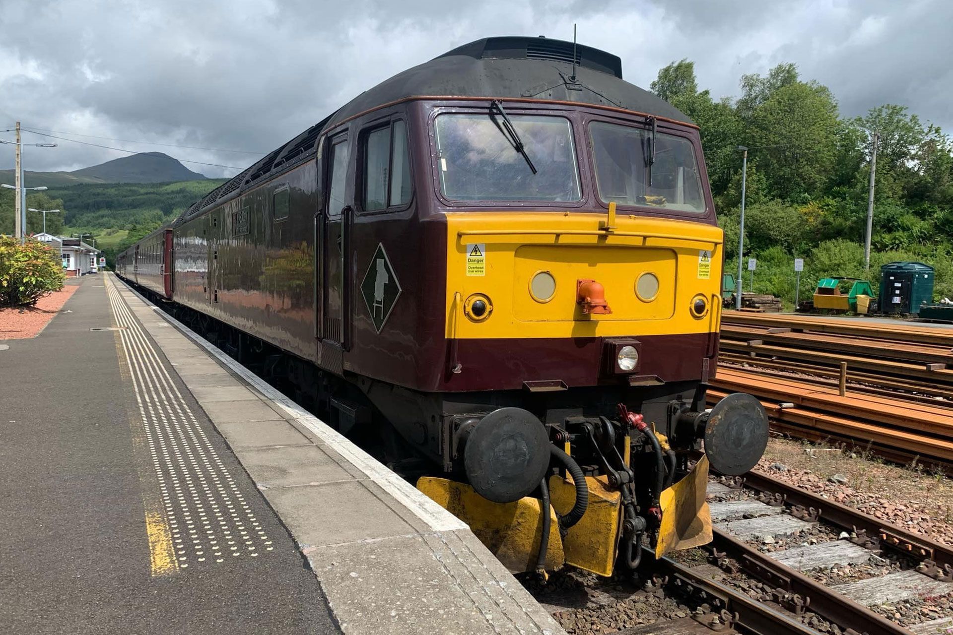 Maroon and yellow diesel locomotive at a platform on a rainy station track, with green hills behind.
