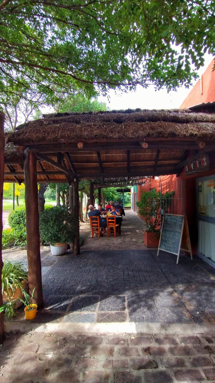 Patio exterior de un restaurante con techo de paja, suelo de ladrillo y gente comiendo en mesas a la sombra.