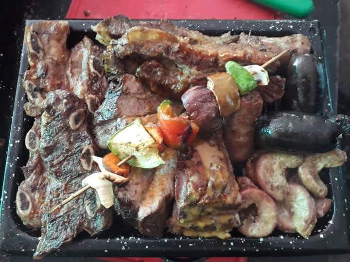 Una bandeja metálica llena de diversas carnes a la parrilla, incluyendo costillas, salchichas y brocheta