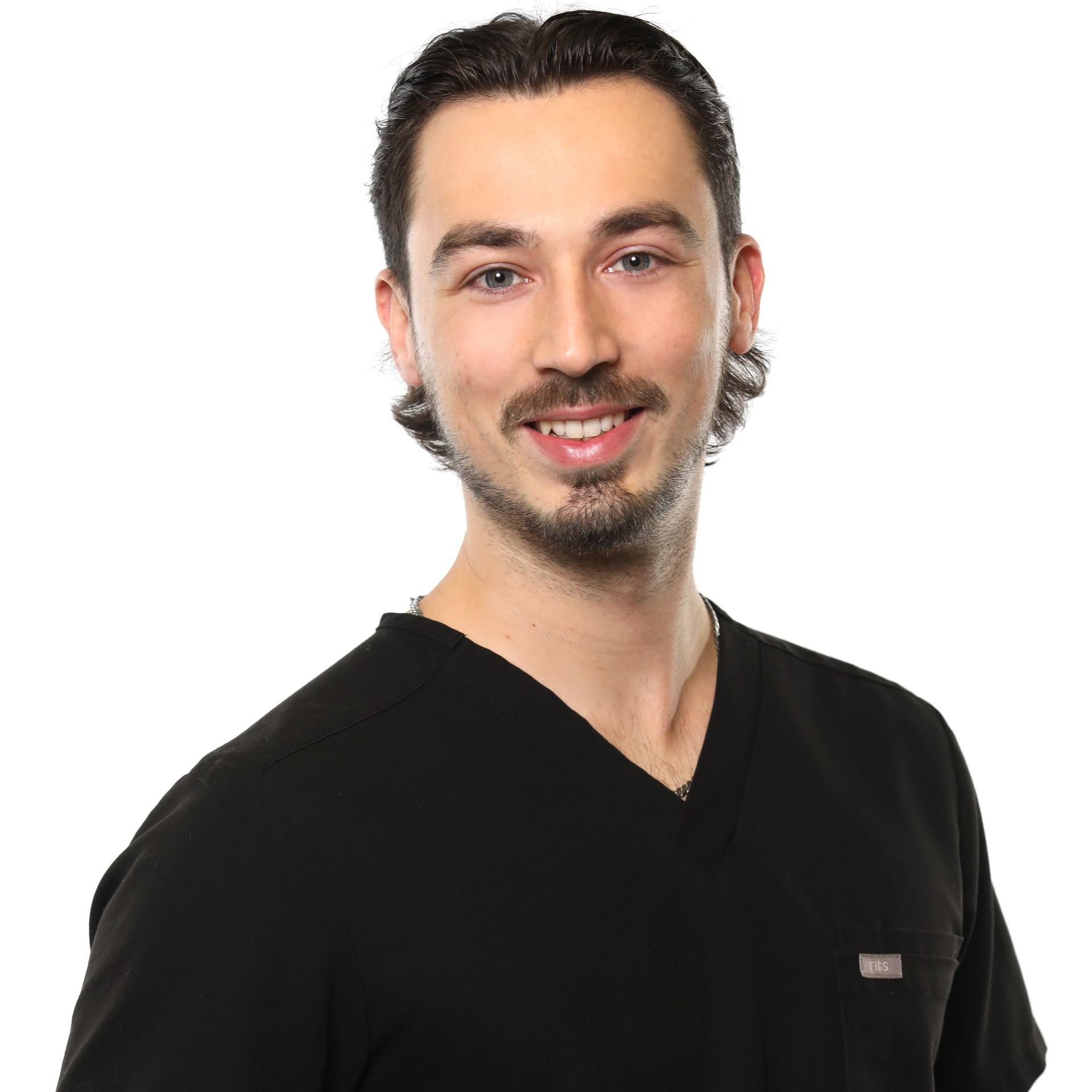 Un homme souriant aux cheveux noirs, à la barbe de trois jours et portant un haut noir sur fond blanc.