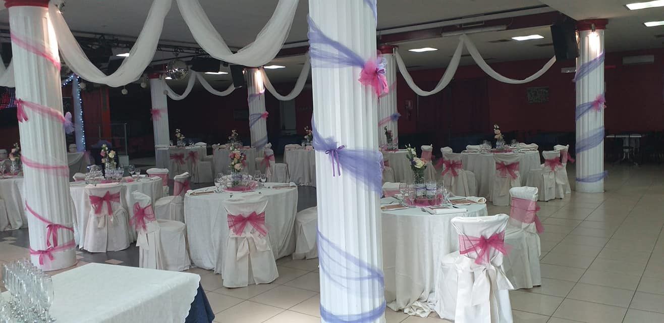 Sala da banchetto decorata con coprisedie rosa, tovaglie bianche e colonne avvolte in tessuto viola.
