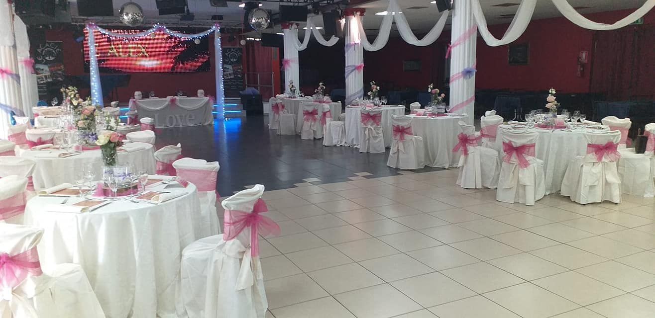 Sala da banchetto decorata con tavoli rotondi, sedie bianche, fasce rosa e un palco illuminato in fondo.