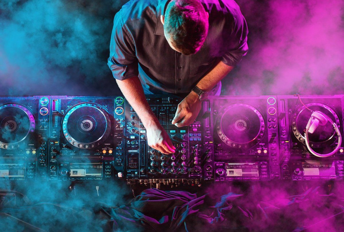 Un DJ mixa su una console illuminata al neon tra fumo rosa e viola.