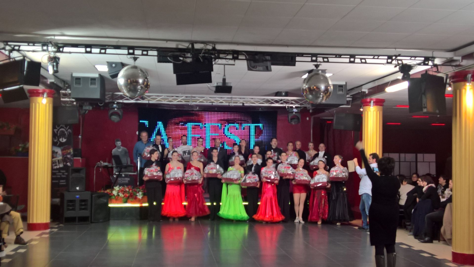 Artisti in costumi colorati su un palco illuminato durante uno spettacolo di danza, con musicisti e pubblico nelle vicinanze.