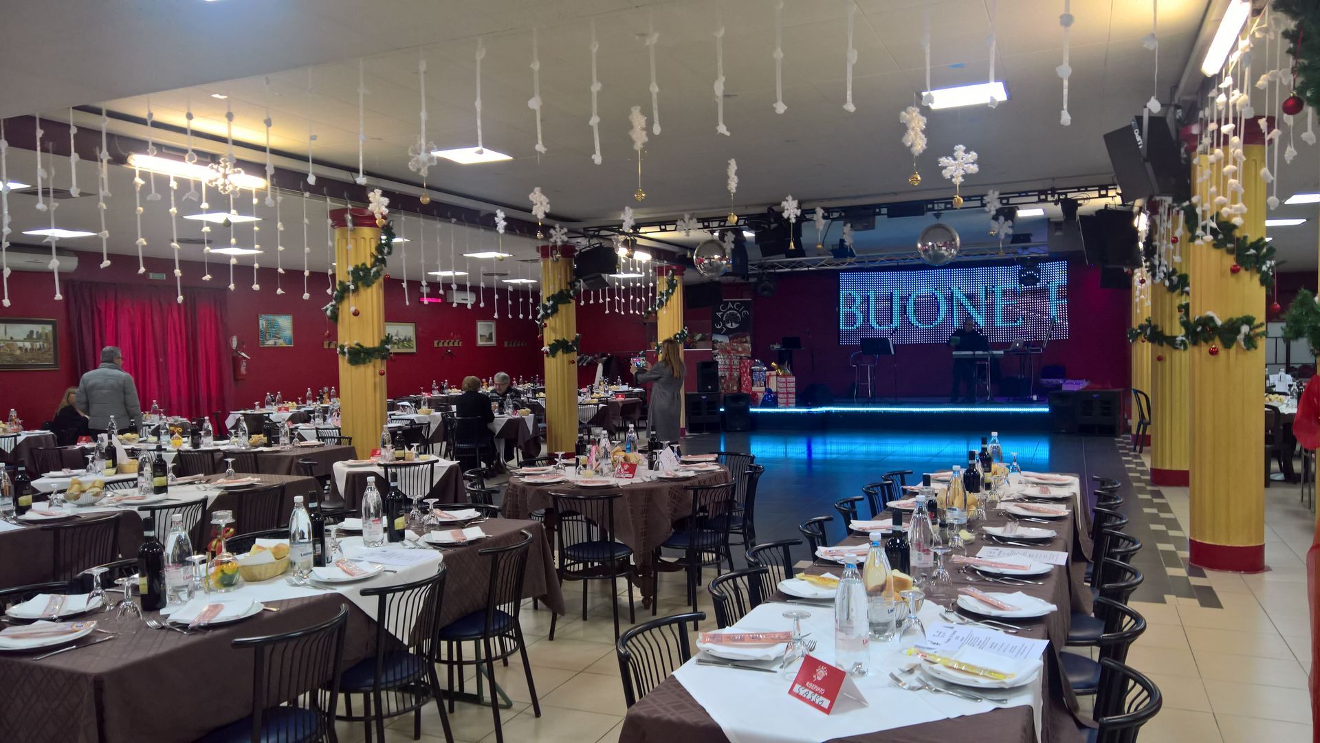 Sala per banchetti con tavoli e sedie apparecchiati di fronte a un palco illuminato con tende blu e rosse.
