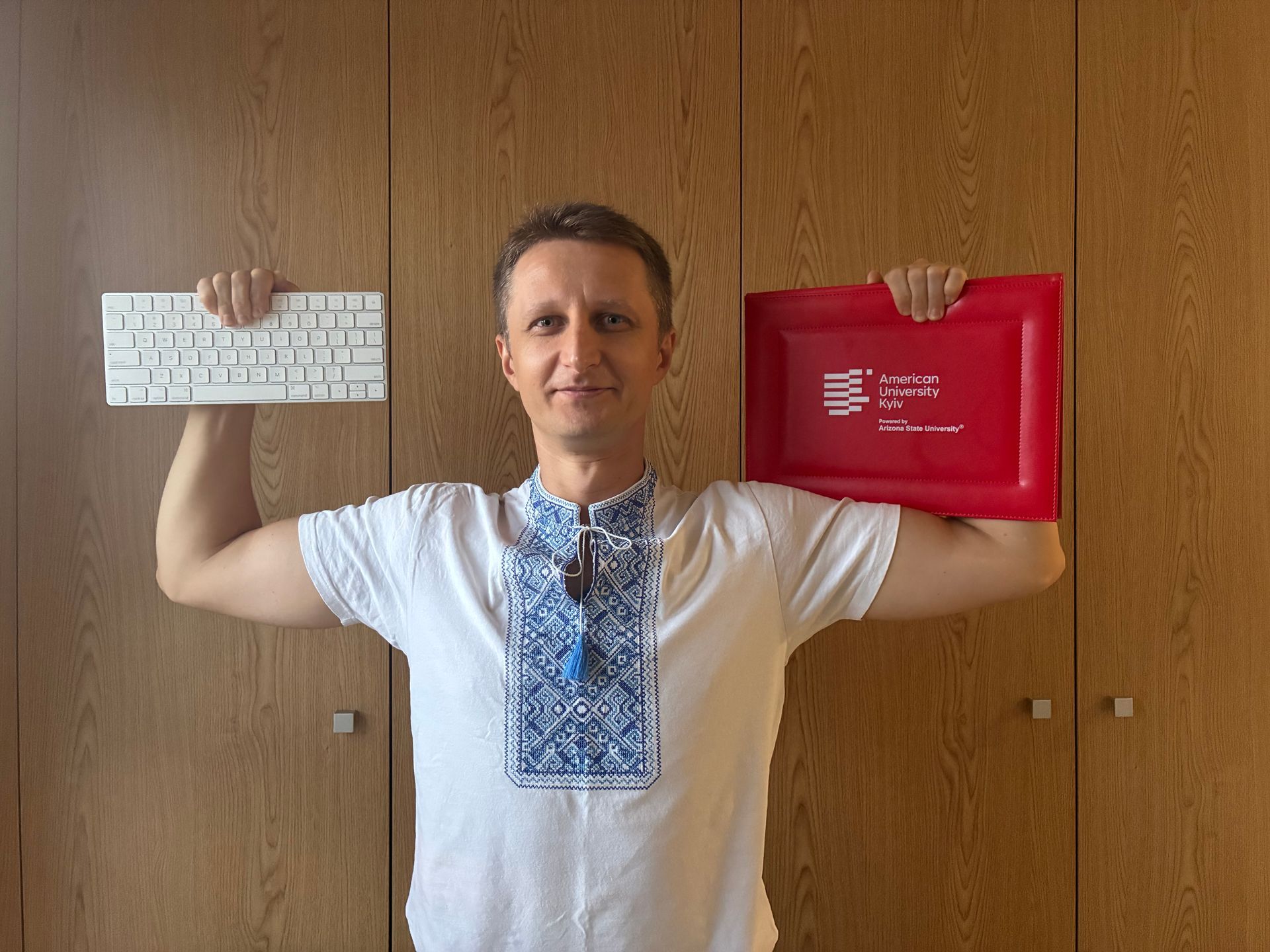 Victor proudly holding a white keyboard and red diploma in front of a wooden wall, wearing a white embroidered shirt
