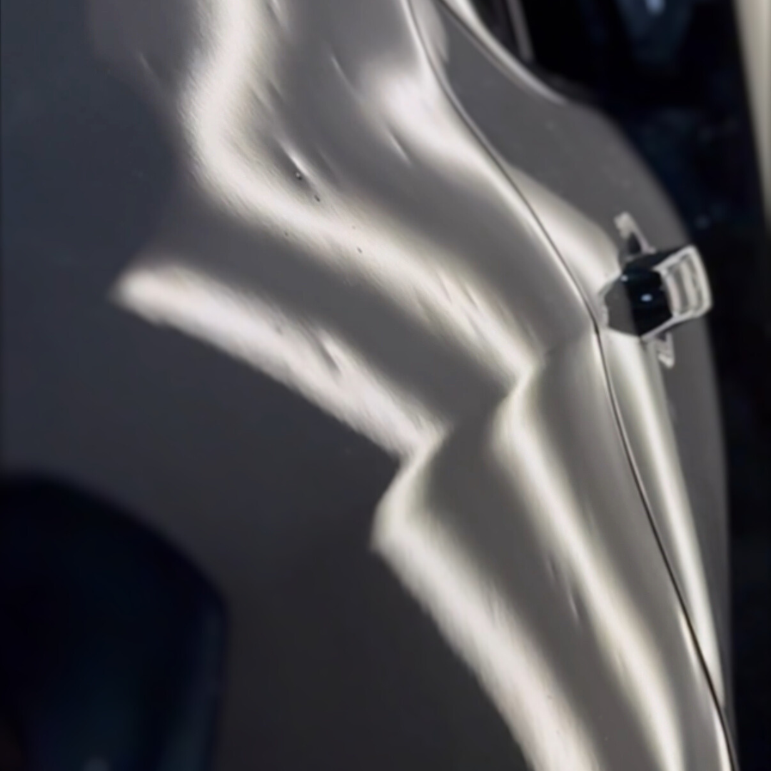 Silver car door with multiple dents, reflecting bright light.