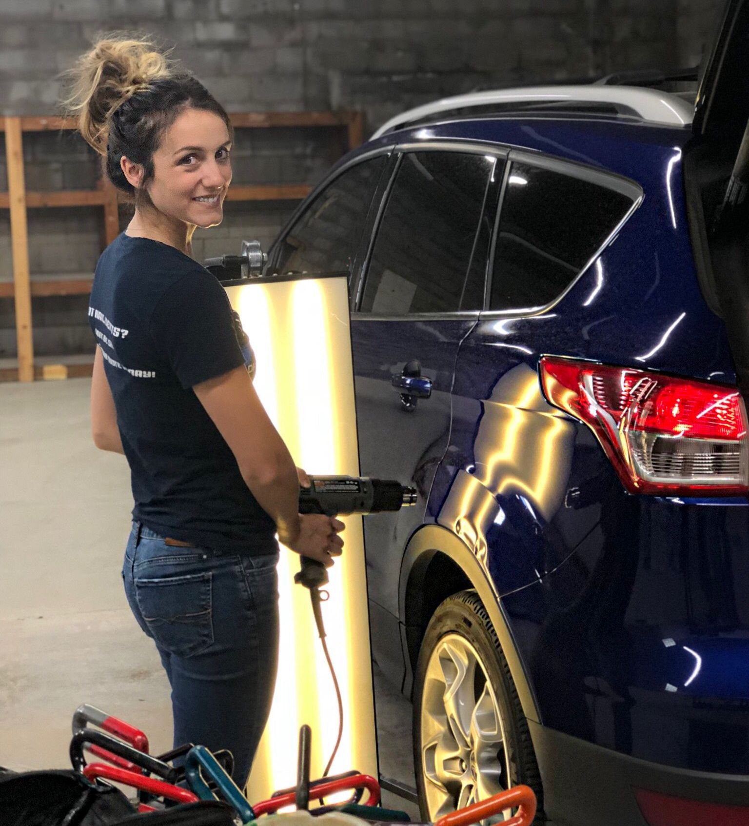 Woman using a tool to repair a dent on a blue car in a garage, smiling.