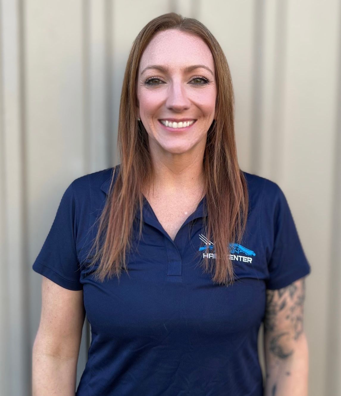 Woman with auburn hair, smiling, wearing a navy blue polo shirt with a company logo, in front of a neutral background.