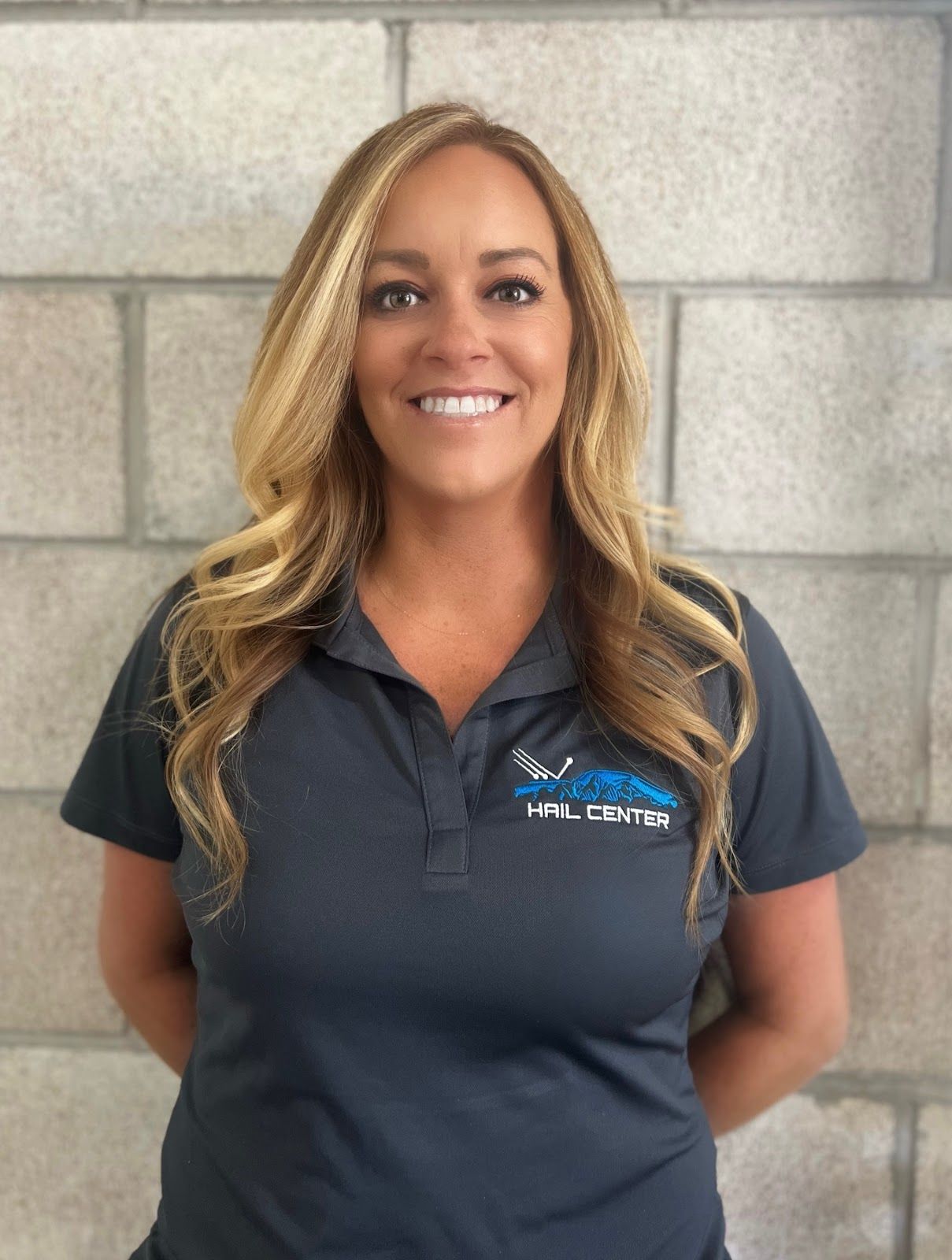 Woman with blonde hair wearing a gray polo shirt, smiling in front of a brick wall.