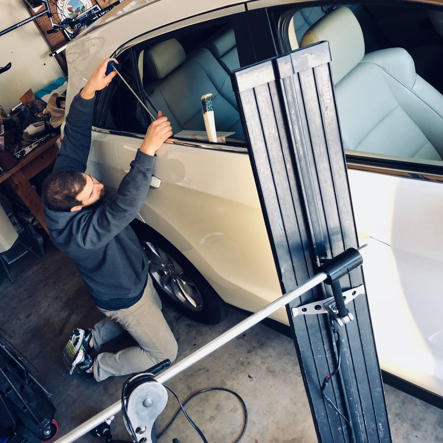 Person kneeling, working on the side window of a white car. He holds a tool near the window.
