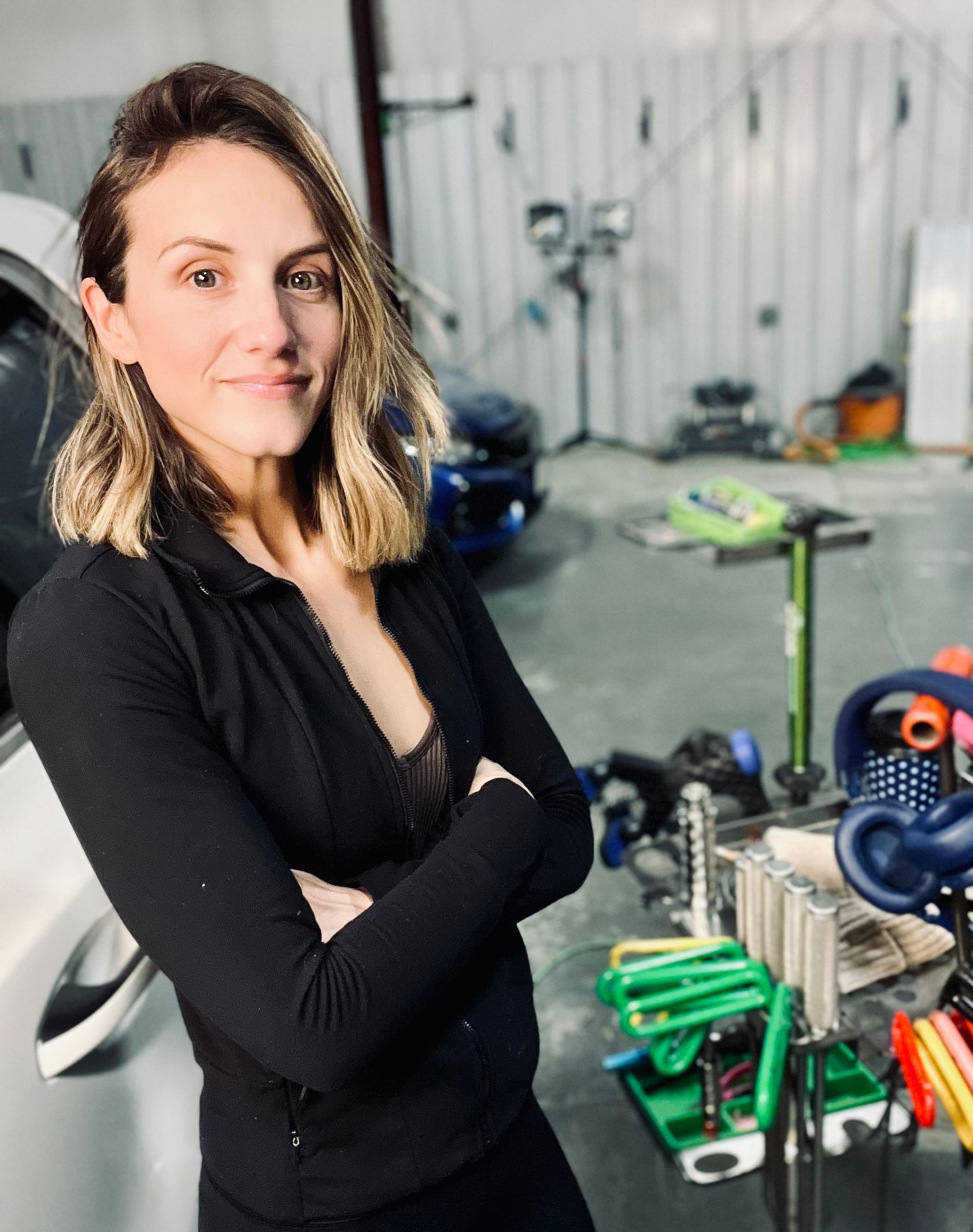 Woman in black standing in a workshop with arms crossed, smiling. Tools and a car are in the background.