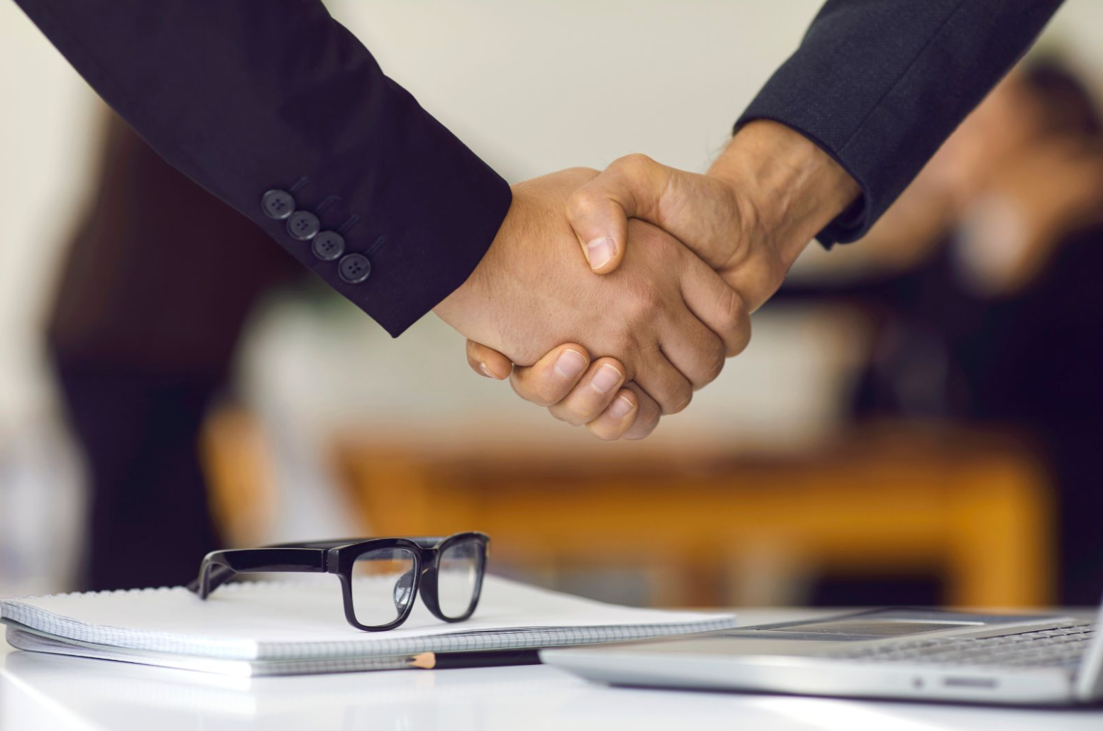Two people shaking hands, sealing a deal; notebook, glasses, and laptop in the foreground.