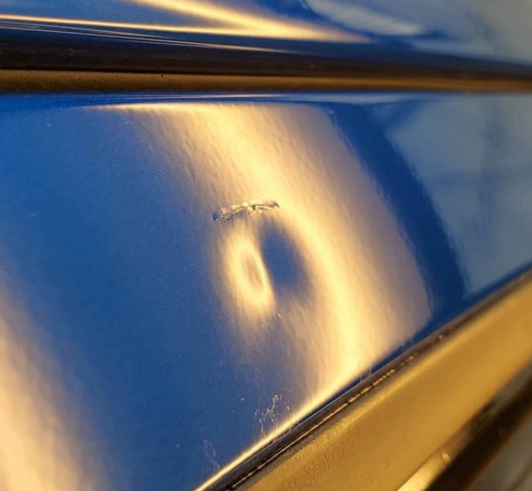Blue car panel with dent and scratches, reflecting bright light.