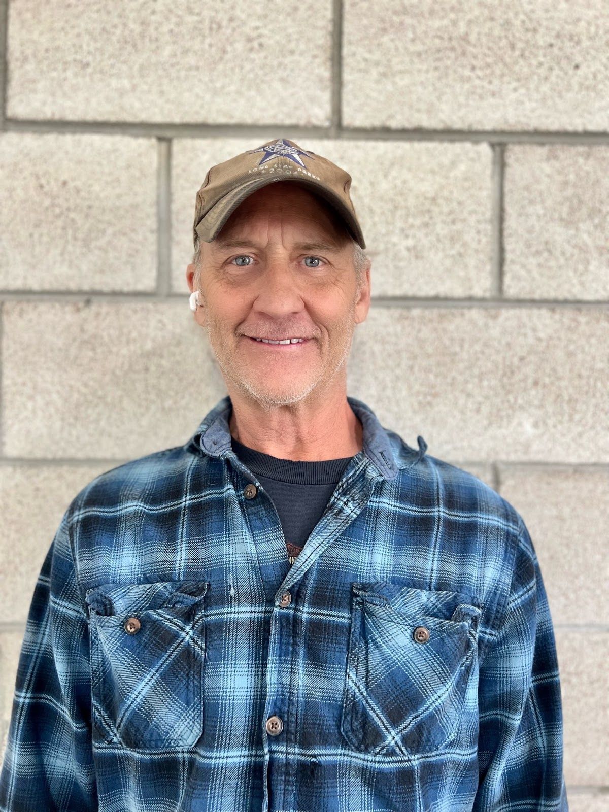Man in blue plaid shirt and cap smiles against a brick wall.