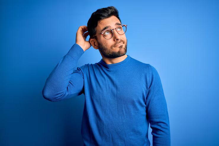 Man with a beard and glasses scratches his head, looking up, against a blue background.