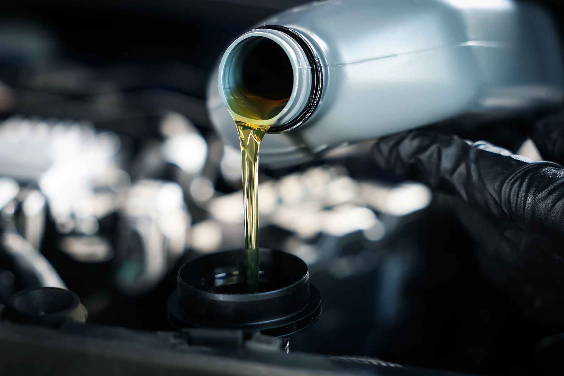 Oil pouring from a silver bottle into an engine fill cap in a car engine bay.