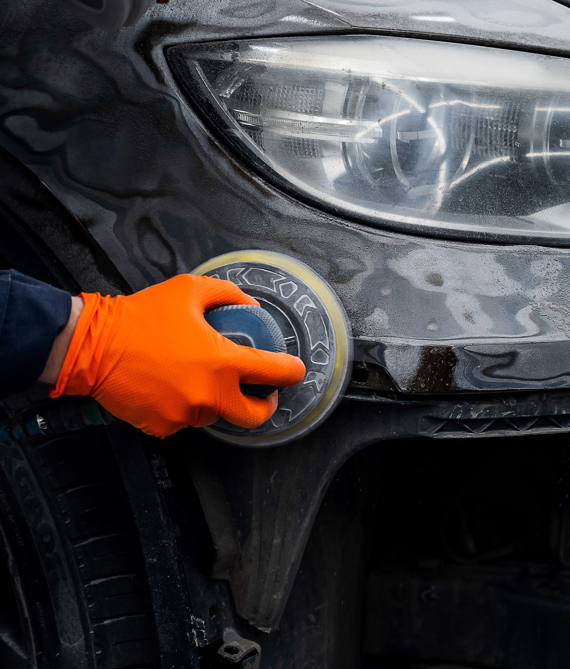 Orange-gloved hand polishing a black car’s headlight with a circular buffer