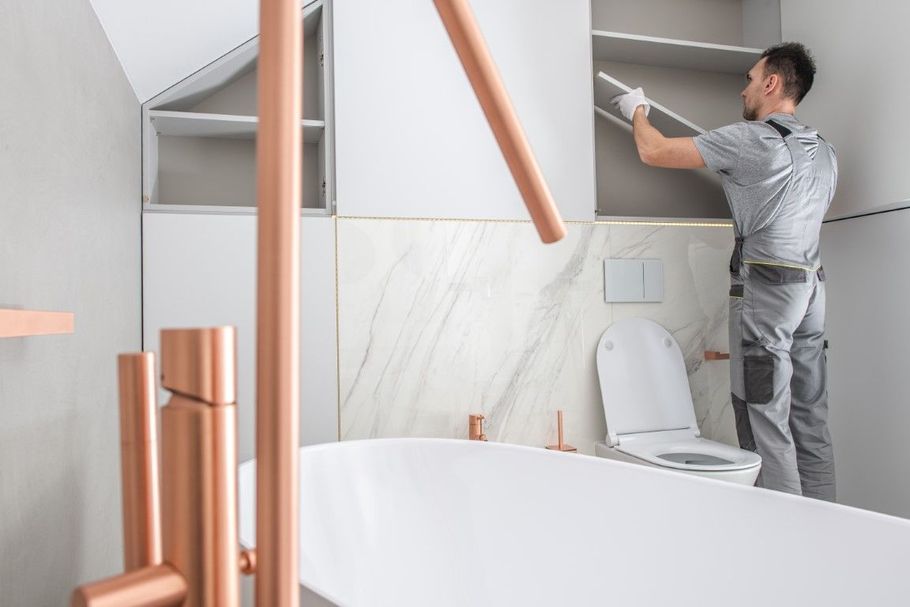 Worker installing a cabinet in a modern bathroom with a toilet, sink, and white marble tiles.