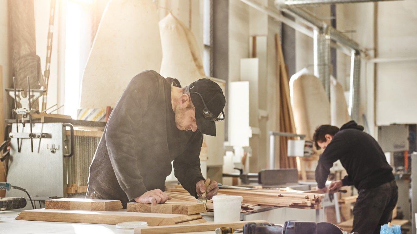 Two people working on wooden boards in a sunlit workshop, surrounded by tools and sawdust