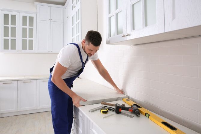Worker in blue overalls measuring a white countertop in a bright kitchen with tools nearby