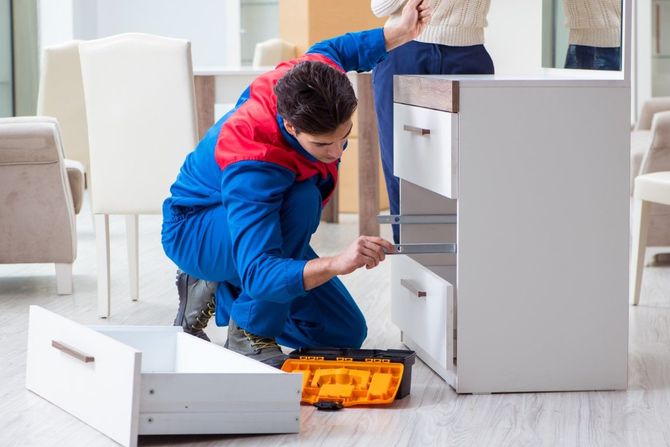 Person kneeling beside white furniture, using tools to fix a drawer in a bright room