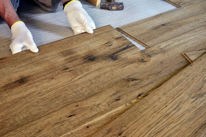 Gloved hands installing a wood plank floor with a utility knife nearby