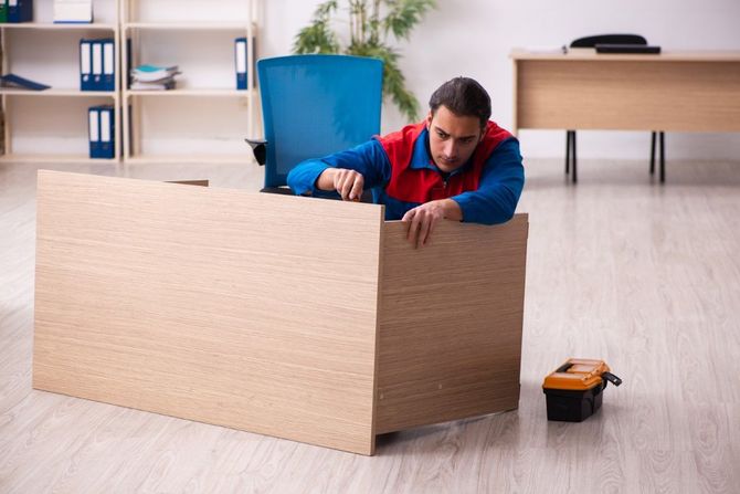 Man in a red-and-blue shirt assembling a large cardboard box in a bright room