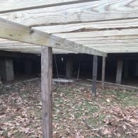 The area beneath a raised wooden deck, showing support posts, joists, and a patch of ground covered in fallen leaves.