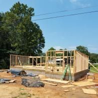 A wooden frame of a house under construction on a concrete foundation, set in a sunny, rural outdoor area with trees.