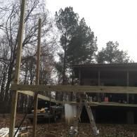 A wooden frame for a deck or platform stands in a wooded yard with an existing house structure in the background.