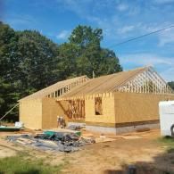 A wooden house under construction, with exposed framing and plywood walls, set against a background of trees and blue sky.