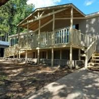 A new wooden deck and porch cover attached to a single-story home, viewed from a dirt and concrete driveway.