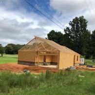 A wooden house frame under construction sits on a dirt lot near a grassy field and trees under a cloudy sky.