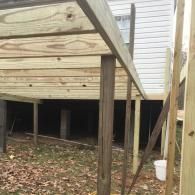 A partially constructed wooden deck frame elevated on posts against the exterior wall of a house, seen from the yard.