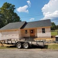 A single-story house under construction with light-colored siding and dark shingle roofing, parked next to a utility trailer.
